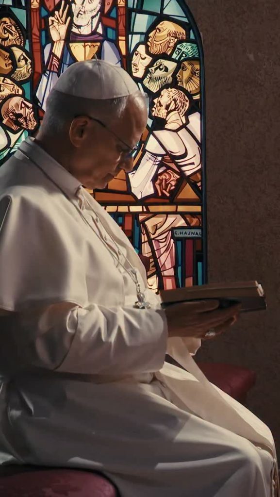 Pope Leo XIV dressed in white bows his head in prayer before a colorful stained-glass window, holding a prayer book
