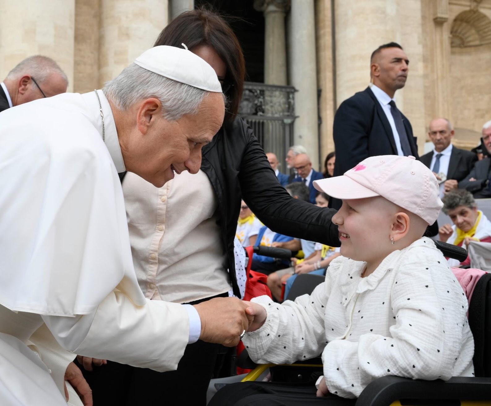 Pope Leo XIV bends down smiling and holds the hand of a young girl in a pink cap seated in a wheelchair at St. Peter's Square